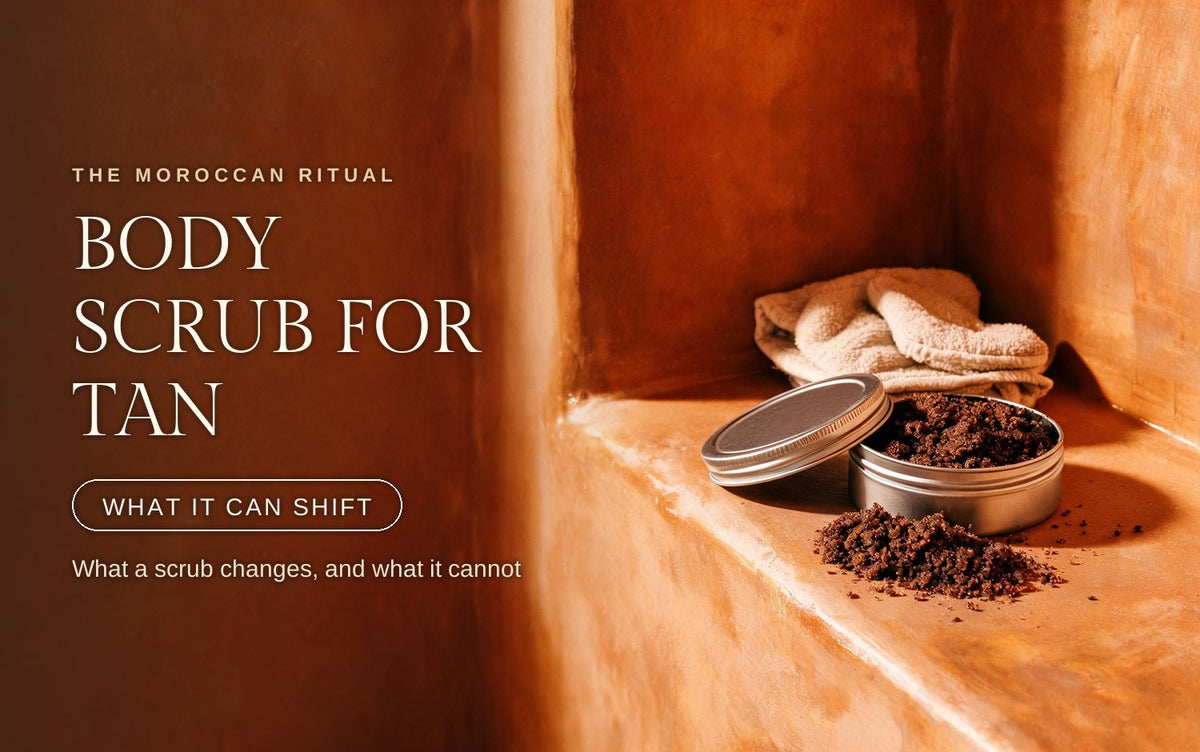 Open steel tin of dark coarse oil based scrub on a warm clay ledge, granules spilled across the stone, a folded terry kessa mitt resting behind it in raking amber light