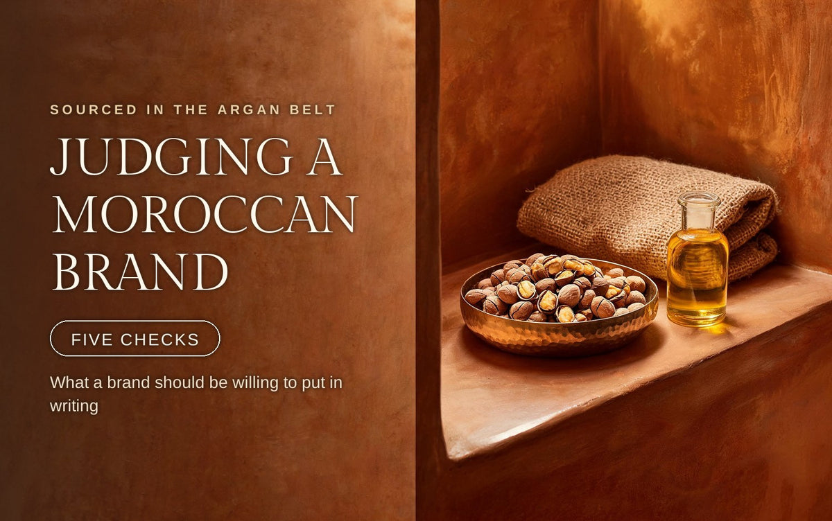 Whole cracked argan kernels heaped in a shallow hammered brass dish beside a small clear glass vial of golden argan oil, a coarse jute sack folded behind them on a warm terracotta tadelakt ledge