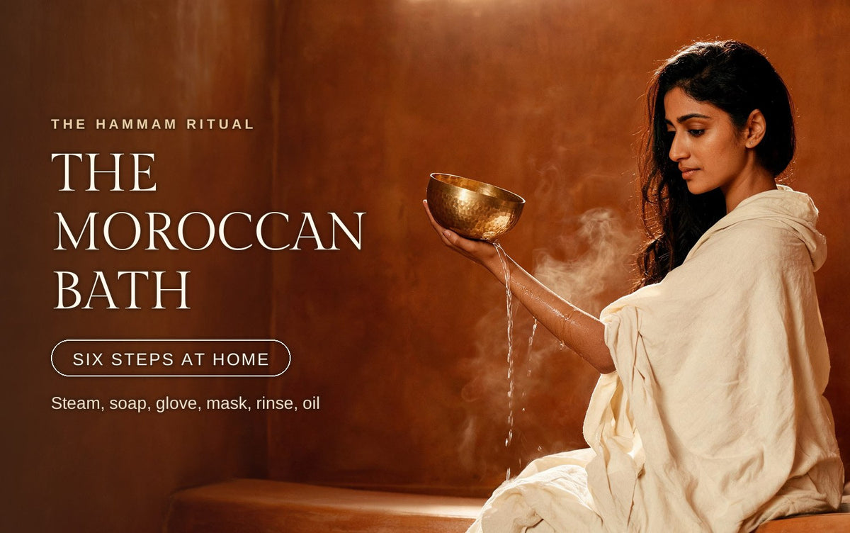 A woman in a cream linen robe pouring water from a hammered brass bowl over her forearm in a steamy Moroccan hammam