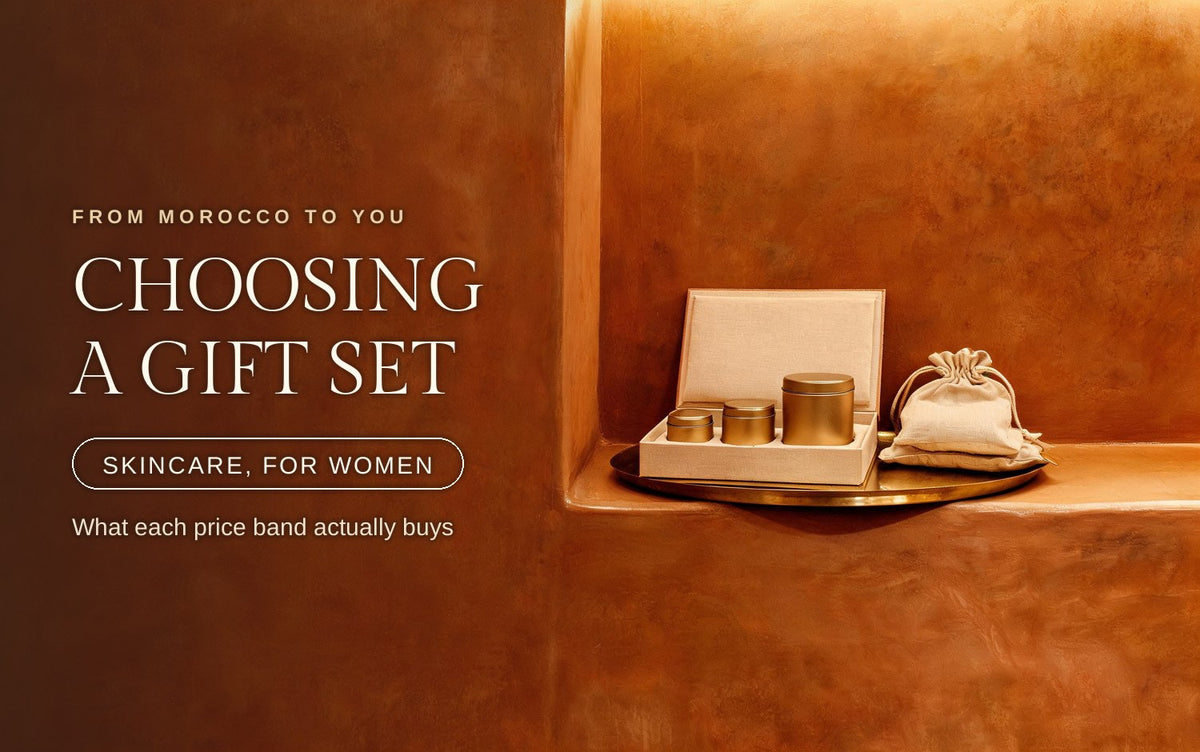 An open gift box lined with pale linen holding three matte gold tins in ascending sizes, a gathered linen drawstring pouch resting beside it on an oval brass tray in a warm terracotta plaster niche